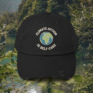 May include: Black distressed baseball cap with the text "CLIMATE ACTION IS SELF-CARE" surrounding a graphic of the Earth. The cap has a curved brim and a distressed look.