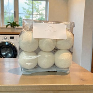 May include: A clear plastic bag containing six white wool dryer balls. A white label on the bag reads "Ocean Wool Dryer Balls". The balls are spherical and appear to be made of a soft, textured material. The bag is sitting on a wooden surface.