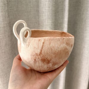May include: A terracotta-coloured ceramic bowl with a white, looped handle. The bowl has a textured surface with white paint strokes. The bowl is held in a person's hand. The bowl is likely used for decorative purposes.