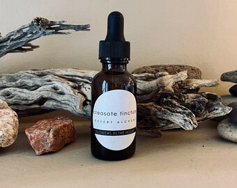 Può includere: Un piccolo flacone contagocce in vetro ambrato con tappo e bulbo in gomma nera. L'etichetta circolare bianca riporta il testo "creosote tincture", "DESERT ALCHEMY" e "FLOWERS IN THE HOUSE" stampato in nero sulla parte anteriore.