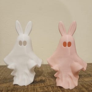 May include: Two ceramic ghost figurines with bunny ears. One is white and the other is pink.