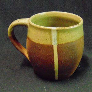 May include: A ceramic mug with a rounded body and a single handle. The upper portion and a vertical stripe running down the front are finished in a light yellow-green glaze, while the lower body features a textured, dark reddish-brown finish.