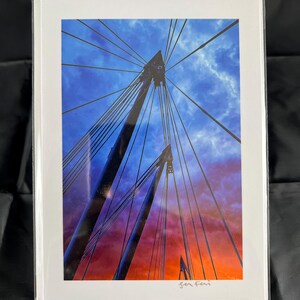 Puede incluir: Una fotografía en blanco y negro de un puente con un cielo azul y naranja de fondo. El puente está hecho de cables de acero y torres. La fotografía está firmada "Jen Fei" en la esquina inferior derecha.