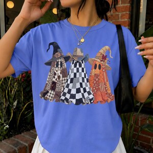 May include: A periwinkle blue short-sleeve t-shirt featuring a graphic print of three ghosts wearing witch hats. The ghosts have distinct patterns including leopard print, black and white checkered squares, and a floral orange autumn design.