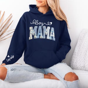May include: Navy blue hoodie with the words "Boy Mama" in white script and patchwork letters. The letters are a mix of blue gingham and floral patterns. The sleeve has the name "Camilo" in white script.