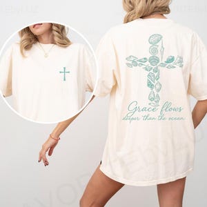 May include: Cream-colored t-shirt featuring a teal cross design on the front and back. The back design is a cross made of seashells, with the text "Grace flows deeper than the ocean" below it. The front of the shirt has a small teal cross.