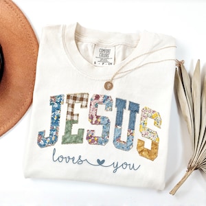 May include: Cream-colored t-shirt with the word "JESUS" in patchwork letters, each with a different floral or plaid pattern. Below, the text reads "loves you" with a heart symbol. A gold necklace and a brown hat are visible.