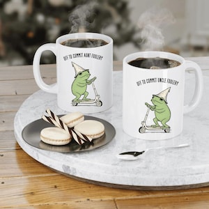 May include: Two white ceramic mugs with black coffee. Each mug features a green frog riding a scooter and wearing a pointed hat. Text on the mugs reads "OFF TO COMMIT AUNT FOOLERY" and "OFF TO COMMIT UNCLE FOOLERY".