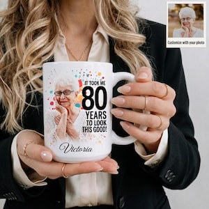 May include: A white ceramic mug featuring a personalized photograph of an elderly person with glasses, the text IT TOOK ME 80 YEARS TO LOOK THIS GOOD!, and the name Victoria in script font, accented by colorful paint splatters on the front surface.