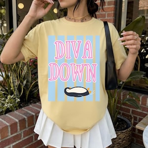 May include: A yellow t-shirt with the words "DIVA DOWN" in pink outlined in white, set against a light blue striped background. A cartoon penguin is below the text. The shirt is a casual style.