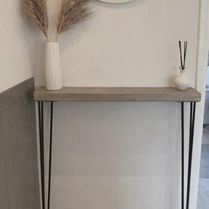 May include: A grey wooden console table with black hairpin legs. A white vase with dried plants sits on the left, and a small white vase with reeds is on the right. A round mirror hangs on the wall above the table.