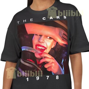 May include: Black t-shirt featuring a graphic of a woman with red lipstick and a hand covering her eye. The text "THE CARS" is above the image, and "1978" is below. The shirt is a vintage style.