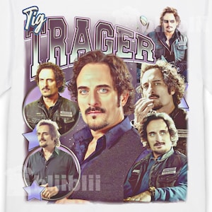 May include: White t-shirt featuring a collage of images of a man with dark hair and a mustache. The name "Tig Trager" is printed in a purple and white stylized font at the top of the design. The design has a vintage aesthetic.
