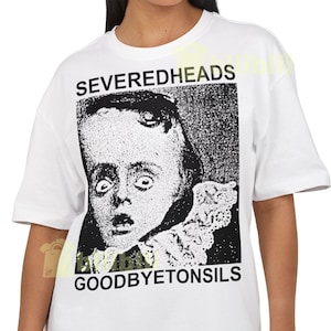May include: White t-shirt featuring a black and white graphic of a person with a surprised expression. The text "SEVEREDHEADS" is above the image, and "GOODBYETONSILLS" is below. The shirt has short sleeves.