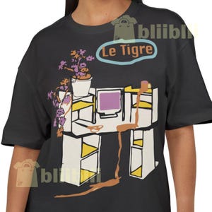 May include: Dark grey t-shirt with a graphic of an office desk, computer, plant, and coffee cup. The text "Le Tigre" is in a blue oval above the desk.