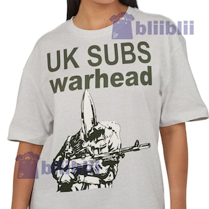 May include: A light gray t-shirt with the text "UK SUBS warhead" in dark green. Below the text is a black and white graphic of a hand holding a rifle. The t-shirt has short sleeves and a crew neck.