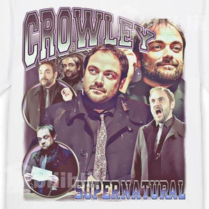May include: White t-shirt with a collage of images of a man in a black coat and tie. The word "CROWLEY" is printed in a purple and white font above the images, and "SUPERNATURAL" is printed below.