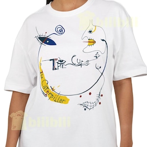 May include: White t-shirt featuring a colorful abstract design with the text "The Cure" and "Caterpillar". The design includes blue, yellow, and black lines and shapes, creating a unique and artistic look. The shirt has short sleeves and a crew neck.