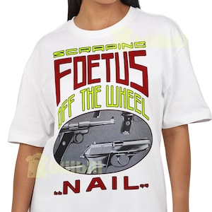 Puede incluir: Camiseta blanca con un diseño gráfico. El texto dice "SCRAPING FOETUS OFF THE WHEEL" en rojo y amarillo. El diseño incluye ilustraciones de pistolas y la palabra "NAIL" en la parte inferior.