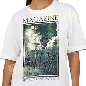 Peut inclure: T-shirt blanc avec le mot "MAGAZINE" imprimé en haut. Le t-shirt présente un graphisme vert et bleu avec les mots "SECONDHAND DAYLIGHT" en bas. Le graphisme a un aspect vintage et vieilli.