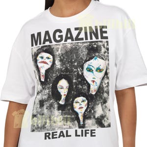 Peut inclure: T-shirt blanc avec le mot "MAGAZINE" au-dessus d'un graphisme de quatre visages stylisés. Le graphisme est dans des tons de gris et de noir, avec les mots "REAL LIFE" en dessous.