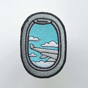 May include: Embroidered patch depicting an airplane window view. The design shows a light blue sky, white clouds, and part of an airplane wing. The patch has a grey border and a black outline.