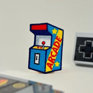 May include: A blue, yellow, and red arcade machine patch with the word "ARCADE" on the side. A grey Nintendo controller and game cartridge are in the background. The patch has a retro video game theme.