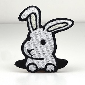 Può includere: Toppa ricamata raffigurante un coniglio grigio che esce da un buco nero. Il coniglio ha l'interno delle orecchie bianco e un contorno nero. La toppa è progettata con uno stile minimalista, adatta per abbigliamento o accessori.