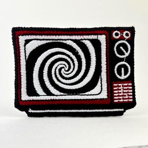 Peut inclure: Écusson brodé représentant un téléviseur rétro. Le téléviseur a un cadre noir avec un bord rouge et un motif en spirale noir et blanc sur l'écran. Le patch est noir avec des coutures blanches.