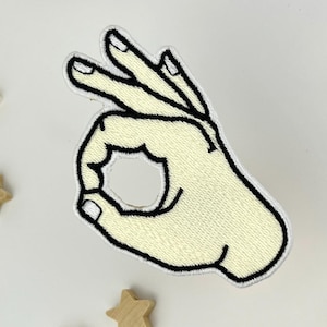 Puede incluir: Un parche bordado de color crema con forma de mano haciendo el gesto "OK", con costuras negras. El parche está rodeado de pequeñas formas de estrella de madera y dos objetos circulares marrones sobre un fondo blanco.
