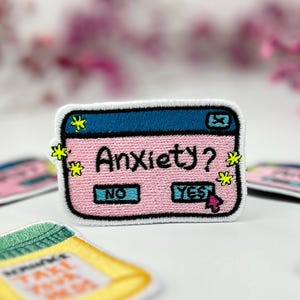 Może przedstawiać: Haftowane naszywki z różowym i niebieskim wzorem okna komputera z napisem "Anxiety?" z przyciskami "No" i "Yes". Inne naszywki mówią "Take Your Meds". Różowe kwiaty w tle.
