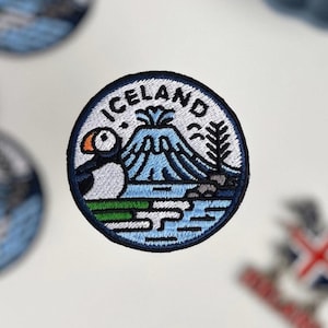 Op de afbeelding: Geborduurde ronde patches met een IJsland-thema. De patches tonen een papegaaiduiker, een vulkaan en het woord "ICELAND". De patches zijn blauw, wit, groen en zwart. Een kopje koffie staat op de achtergrond.