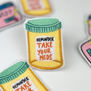 May include: Embroidered patches featuring a yellow pill bottle with a green lid and the text "REMINDER TAKE YOUR MEDS." Other patches include a pink and blue design with the text "Anxiety?" and "Panic53?" on a white background.