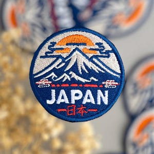 Peut inclure: Écussons brodés ronds avec un motif de montagne bleu, blanc et orange, et le mot "JAPAN" en blanc. Du texte japonais est également inclus. Les écussons ont une bordure bleue.