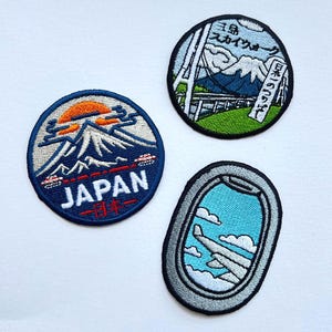 May include: Three embroidered patches with travel themes. One patch features a mountain scene with the word "JAPAN" and Japanese text. Another shows a mountain landscape with Japanese text. The third patch depicts an airplane window with clouds and a wing.