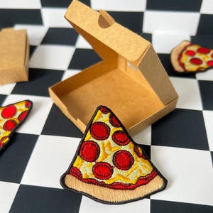May include: Embroidered pizza slice patch with red pepperoni, yellow cheese, and a brown crust. The patch is next to a small, open cardboard pizza box and two other pizza slice patches. The background is a black and white checkered pattern.