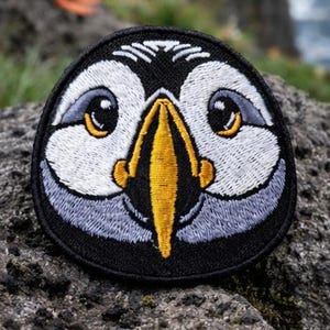 May include: A circular embroidered patch depicting a puffin's face, with a black border, white, grey, and yellow details. A real puffin stands in the background on a rock near the sea.