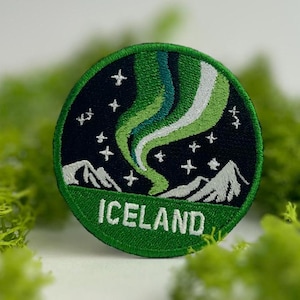 Puede incluir: Un parche bordado redondo con la palabra "ICELAND" debajo de una representación de la Aurora Boreal. El diseño incluye auroras boreales verdes, blancas y azules sobre un cielo nocturno azul oscuro con montañas y estrellas blancas.
