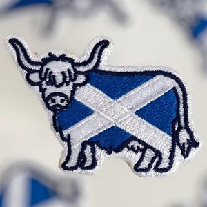May include: Embroidered patch featuring a Highland cow with the Scottish flag design. The cow is blue with a white cross, and the patch has a white border. The background includes blurred patches with the same design.