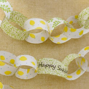 Puede incluir: Guirnalda de cadena de papel con patrones alternos de limones amarillos, hojas verdes y puntos azules sobre fondo blanco. Un eslabón dice "Happy Sukko". Decoración festiva.