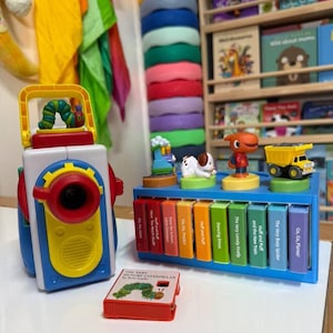 May include: A colorful children's toy set featuring a blue, red, and yellow camera-shaped toy with a caterpillar on top, and a blue toy with miniature book-shaped blocks. A small book titled "The Very Hungry Caterpillar" is also present.