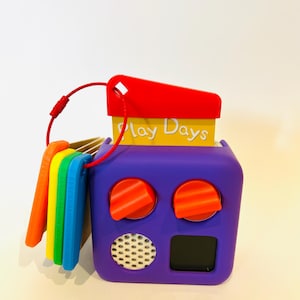 May include: A purple toy with a yellow and orange top that says "Play Days". The toy has two red knobs and a small black screen. There are five colorful plastic keys attached to the side.