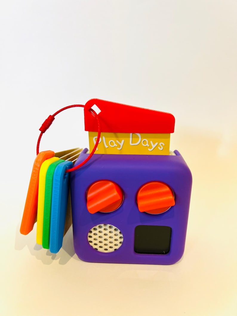 May include: A purple toy with a yellow and orange top that says "Play Days". The toy has two red knobs and a small black screen. There are five colorful plastic keys attached to the side.