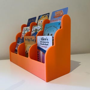 May include: Orange plastic card holder with rounded edges and multiple slots for holding cards. The holder is filled with various cards, including a book cover with the title "Alice's Adventures in Wonderland" and a card with the text "The Ugly Five".