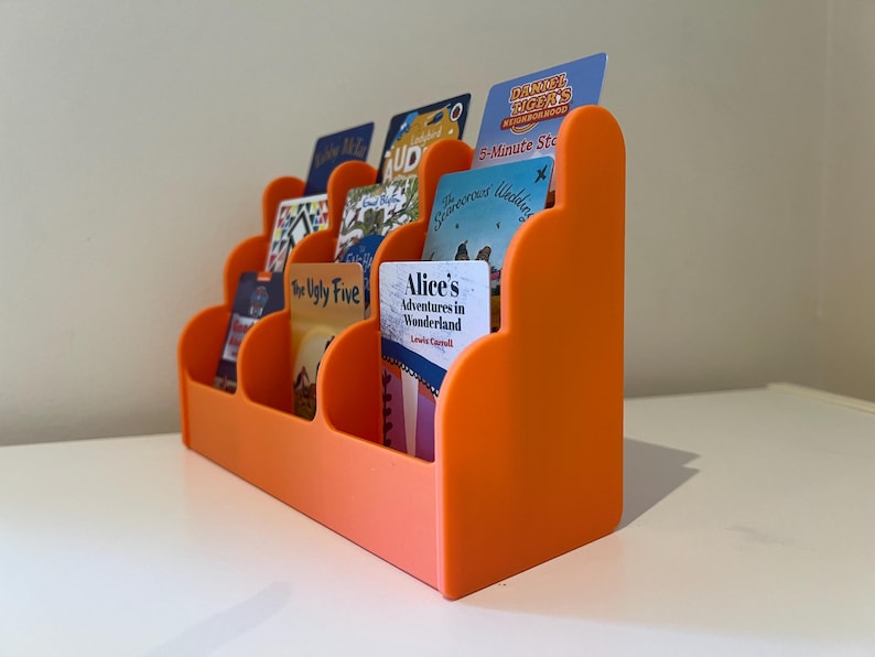 May include: Orange plastic card holder with rounded edges and multiple slots for holding cards. The holder is filled with various cards, including a book cover with the title "Alice's Adventures in Wonderland" and a card with the text "The Ugly Five".