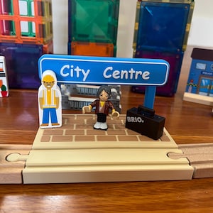 Puede incluir: Un juego de estación de tren de juguete con un letrero azul que dice "City Centre". El juego incluye figuras en miniatura, una maleta negra con "BRIO" impreso y una vía de tren de madera. Azulejos magnéticos de colores en el fondo.