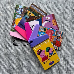 May include: A yellow card with the words "Play Days" and a picture of two cartoon pigs riding scooters. The card is attached to a purple, pink, and white plastic holder with a black string.