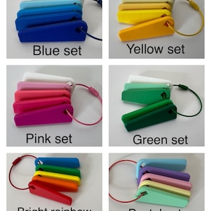 May include: A set of five colorful keychains, each with a different color scheme: blue, yellow, pink, green, and rainbow. The keychains are made of plastic and have a simple, rectangular shape. Each keychain has a small loop at the top for attaching keys.