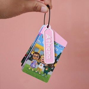 May include: A stack of colorful flashcards featuring cartoon characters, a yellow school bus, and the text "Little Melon Hits". A pink tag with the name "Athema" is attached to the top of the stack.