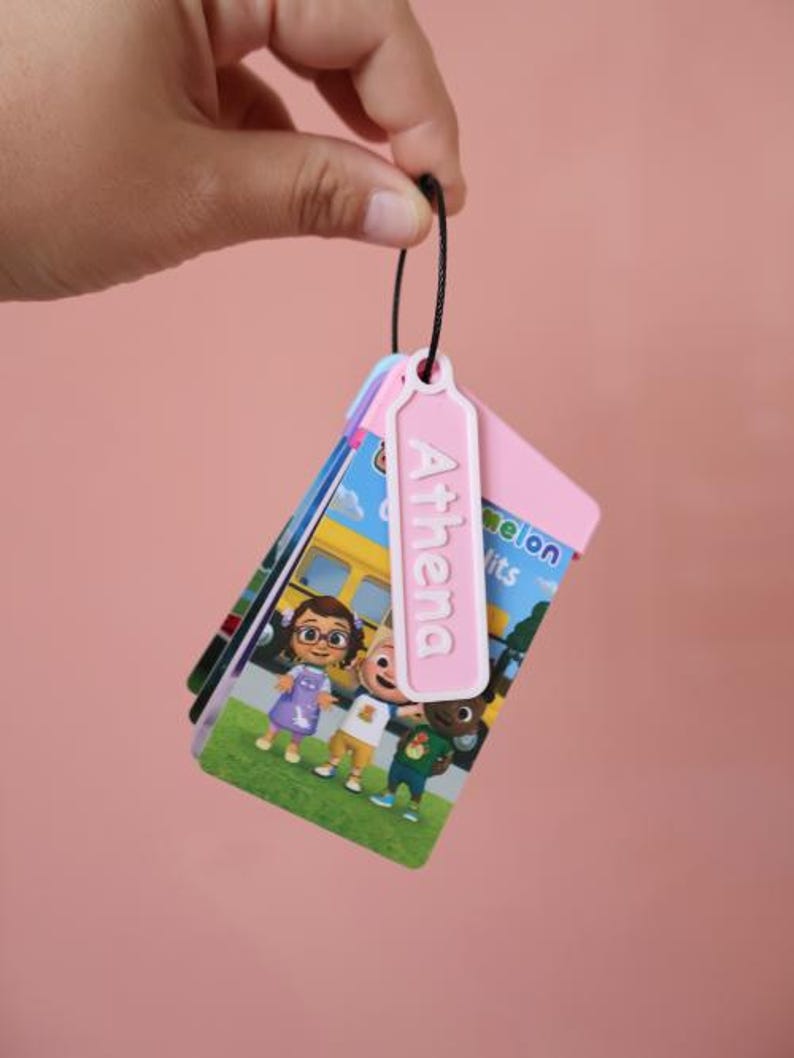 May include: A stack of colorful flashcards featuring cartoon characters, a yellow school bus, and the text "Little Melon Hits". A pink tag with the name "Athema" is attached to the top of the stack.